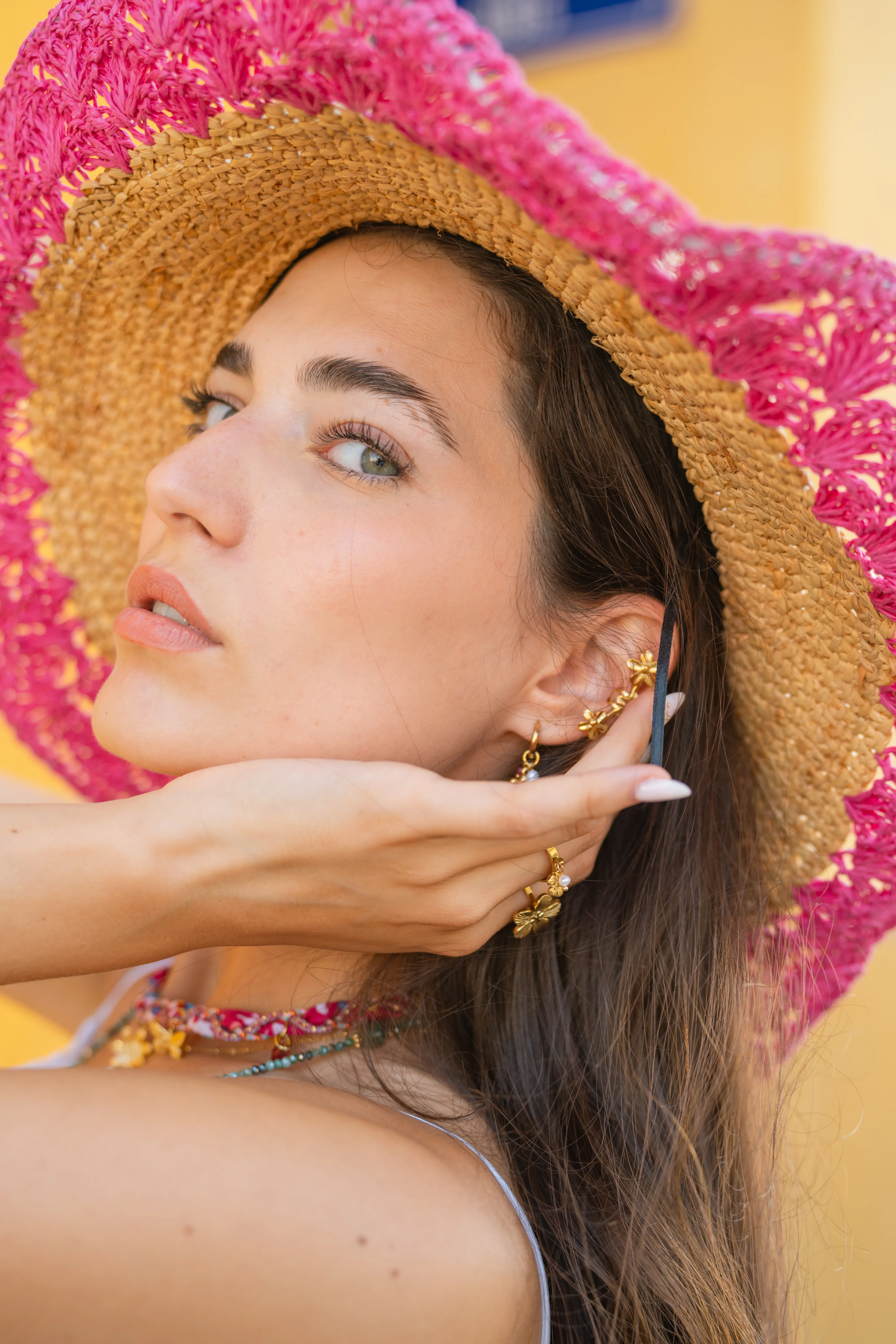 Portrait de profil d'une femme au chapeau de paille fuchsia, geste de la main devant l'oreille avec boucle dorée pendentif fleur Mary Jane