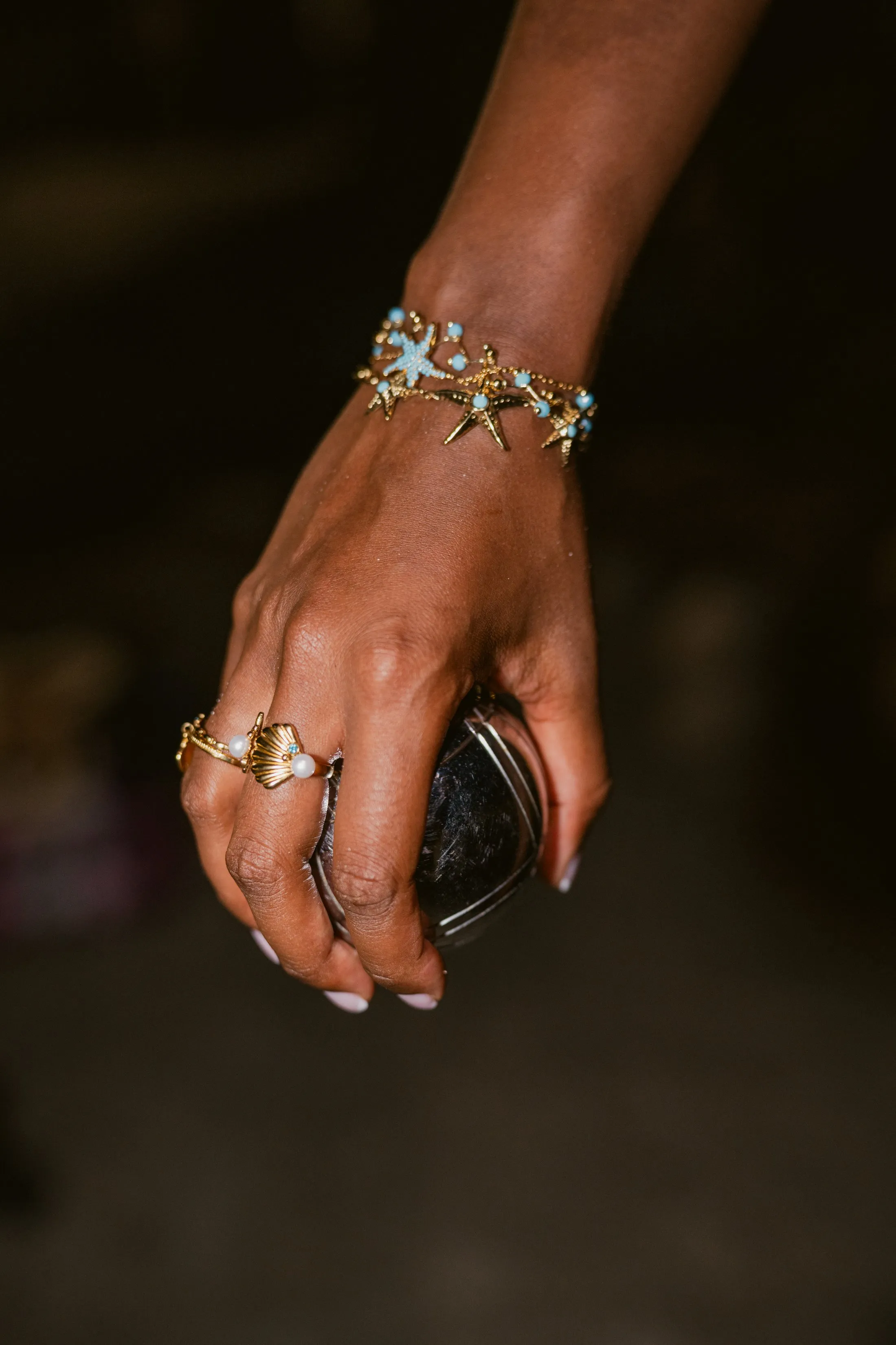Bracelet doré aux étoiles de mer et perles bleues porté au poignet, main sur une boule de pétanque, shooting Mary Jane à Marseille